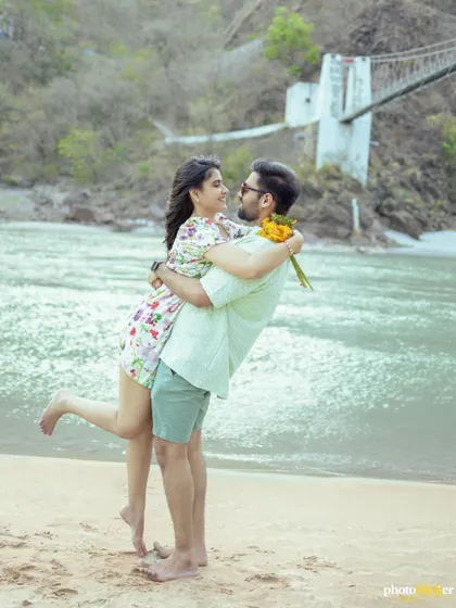 A joyful moment as the groom lifts his bride on a sandy beach in Rishikesh, with the river and a suspension bridge in the background.