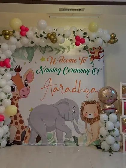 A complete jungle-themed naming ceremony decoration. This setup includes a custom backdrop with cartoon animals, a white and gold balloon arch, and "BABY" letter blocks, creating a perfect spot for photos.