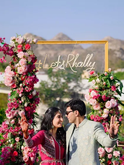 The beautiful couple posing at their mehendi, with a custom neon sign and a backdrop of pink bougainvillea and roses.