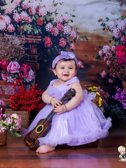 A baby girl in a lovely lavender dress, holding a small guitar in a room full of flowers. The soft colors and her sweet expression make for a very gentle and beautiful portrait.