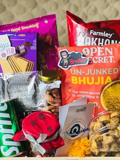 A crate full of savory and sweet snacks for Rakhi, including bhujia, biscuits, and a can of Sprite, decorated with marigold flowers.