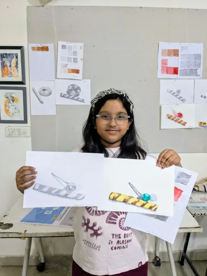 A student proudly displays her pencil and watercolor studies of everyday objects. Notice the value scales in the background, which are exercises we do to master shading.