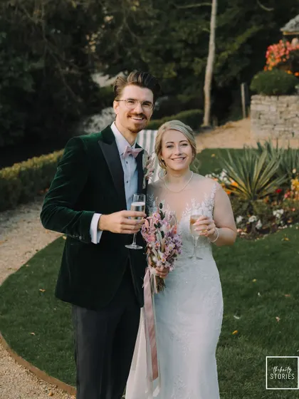 A lovely portrait of the smiling couple, holding their champagne glasses against the lush green backdrop of their garden venue.