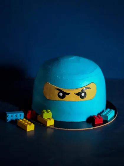 A Lego Ninjago theme birthday cake. The dome shape is carved from cake, and the ninja's face is created with fondant details. The real Lego bricks around the base add a fun, playful touch.