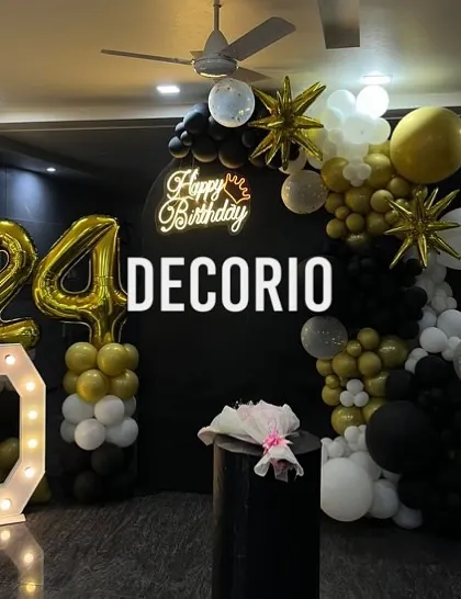 A stylish 24th birthday decoration with a black, gold, and white color scheme. The setup includes a large 'D' marquee letter, '24' foil balloons, and a balloon arch against a black backdrop with a neon sign.