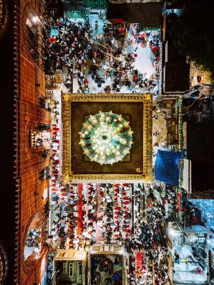 A focused top-down shot of the main tomb at Nizamuddin Dargah, illuminated by a brilliant chandelier. The surrounding crowd and red threads of prayer tied nearby are visible, showing the heart of the devotional activities.