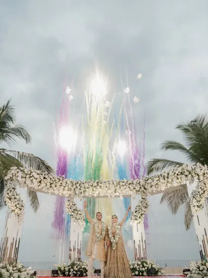 A wide-angle shot showing the scale of the celebration, with colorful fireworks lighting up the sky above the beachside mandap.