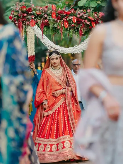 A bride in a stunning red lehenga makes her entrance under a floral canopy held by her loved ones.