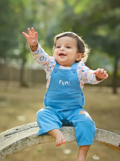 An adorable toddler in blue overalls waves happily during an outdoor photoshoot. The natural setting and the child's joyful expression make for a perfect candid portrait.