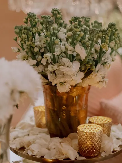 A close-up of the decor, showing simple arrangements of white flowers in textured gold vases, surrounded by candles. The petals scattered on the table add a touch of softness.