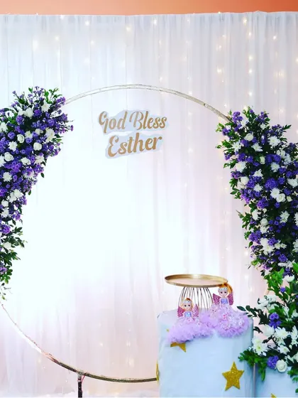 A floral decor setup for the holy baptism of baby Esther. The main feature is a golden ring decorated with beautiful white and purple flowers against a curtain backdrop with fairy lights.