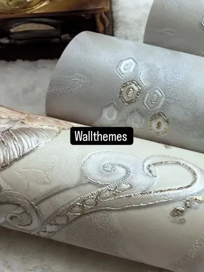 Close-up of textured wallpaper rolls in neutral tones with elegant silver and gold embroidered-look details.