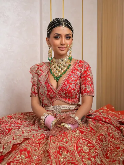 A seated portrait capturing the grandeur and grace of the bride. The rich red lehenga and intricate jewelry are balanced with makeup that is both glamorous and refined, perfect for the main event.