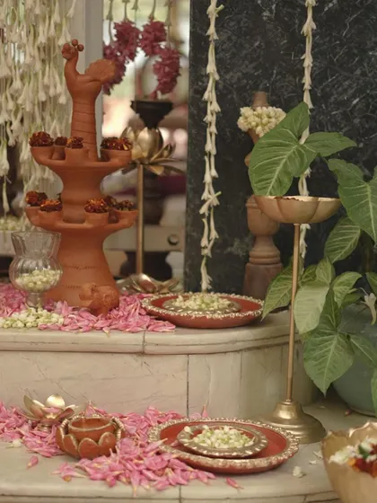 An elegant corner setup for a traditional event, featuring a multi-tiered terracotta lamp and brass plates filled with fresh flowers. The scattered pink petals add a soft, celebratory touch.