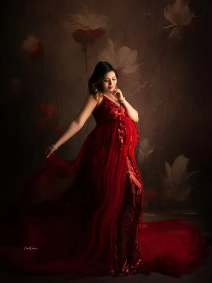 A fine-art maternity portrait with a painterly feel. The mom-to-be poses elegantly in a sequined red gown against a dark floral backdrop.