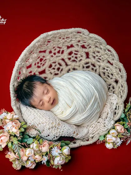 A tightly swaddled newborn in a woven bowl against a bold red background, creating a striking and artistic image.