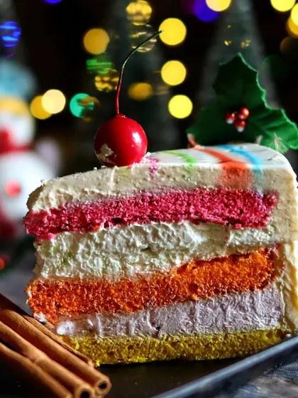 A close-up of the multi-layered rainbow pastry, focusing on the textures of the cake and cream. The cinnamon sticks and holly add a touch of seasonal styling.