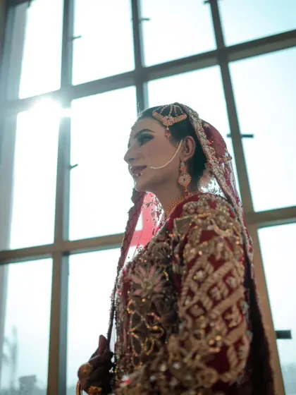 A beautiful, sun-kissed bridal portrait. The light coming through the window creates a soft, ethereal glow around the bride in her intricate red and gold outfit.