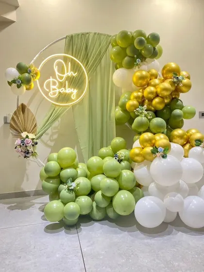 For a chic and modern baby shower, we offer gender-neutral themes like this green and gold setup. It features a circular hoop with an "Oh Baby" neon sign, draped fabric, and an organic balloon garland in olive green, white, and chrome gold.
