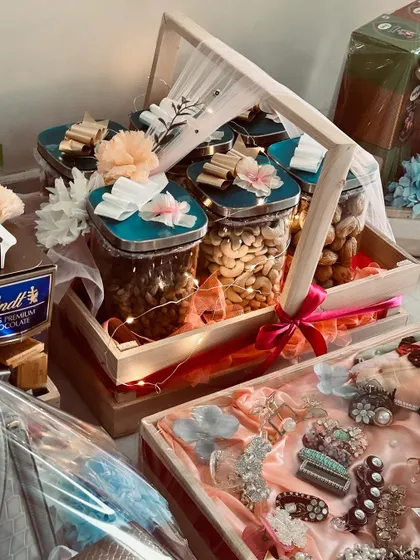 A luxury trousseau hamper featuring jars of premium dry fruits and chocolates. The gifts are arranged in a handled pinewood tray and decorated with flowers and fairy lights.