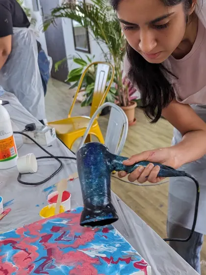 A participant using a hairdryer to manipulate the fluid paints in a pour painting workshop. This technique helps create interesting cells and patterns in the abstract design.