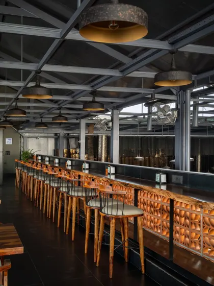 The high-top seating along the side of the space offers a different vantage point. From here, guests can appreciate the soaring ceiling, the natural light from the clerestory windows, and the custom terracotta block wall that adds a warm, earthy element to the bar design.