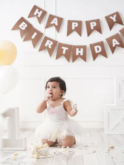A fun shot of a baby girl enjoying her cake smash, with frosting on her face and a 'Happy Birthday' banner in the background.