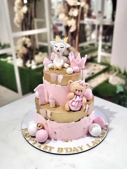 A sweet and elegant three-tier cake for a first birthday, featuring a cute fawn, a teddy bear, and a crown in a lovely pink and gold color scheme.