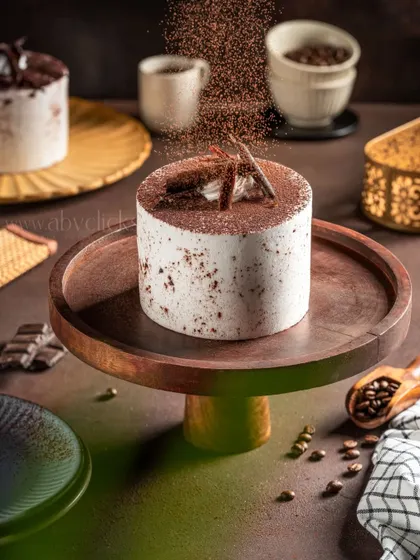 A tiramisu-inspired cake on a wooden stand, with cocoa powder being dusted on top to capture a moment of action and add a sense of freshness.