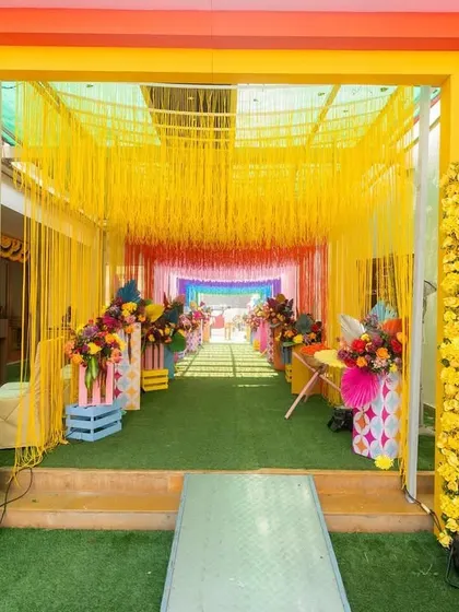 A vibrant tunnel entrance with rainbow-colored floral pillars and a ceiling of yellow tassels, creating a joyful and immersive experience for guests.