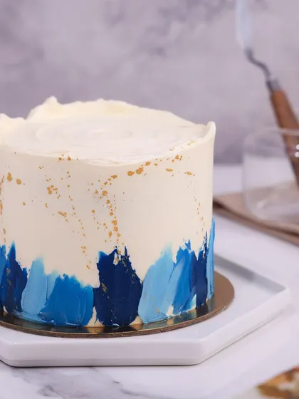 We create a piece of modern art with this abstract blue buttercream cake. Strokes of various blue shades are painted onto a white canvas, accented with gold splatters and topped with piped rosettes, making it a stylish and happy choice for a Father's Day celebration.