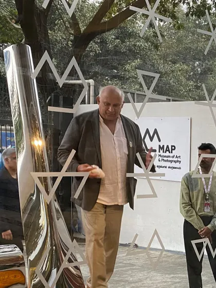 Artist Tarik Currimbhoy speaks beside his polished steel sculpture at the museum's entrance. The reflective surface of the artwork captures the movement of the city, creating a living connection to the urban environment.