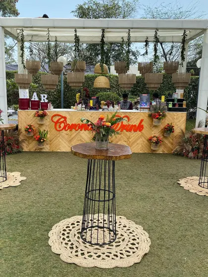 A stylish bar setup for a boho-themed sundowner. The bar front is made of natural wood, complemented by rustic high-top tables and jute decor, creating a relaxed and trendy vibe.