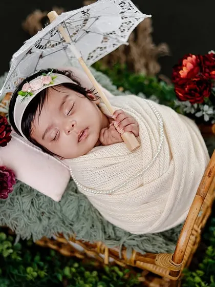A beautifully composed shot of a baby swaddled in a basket, holding a delicate lace parasol and surrounded by greenery and flowers.