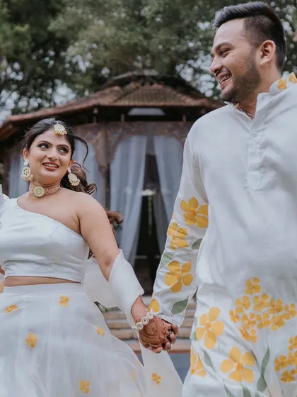 The happy couple, dressed in white and yellow, holding hands and walking through their beautiful Haldi celebration.