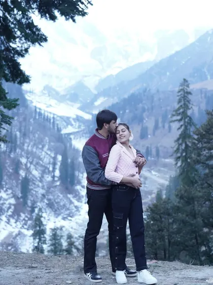 A sweet moment with the groom kissing the bride's forehead, set against a backdrop of snow-covered hills and pine trees in Manali.