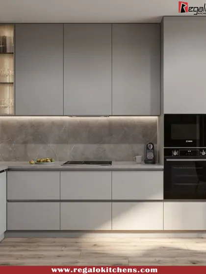 A sleek and minimalist L-shaped kitchen in a uniform grey finish. The handle-less cabinets and integrated appliances create a seamless look. A small, illuminated glass cabinet adds a decorative touch to the functional design.