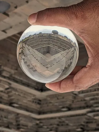 A hand holds a crystal ball that reflects an inverted image of an ancient stepwell. This creative technique offers a unique, mind-bending perspective on architectural photography.