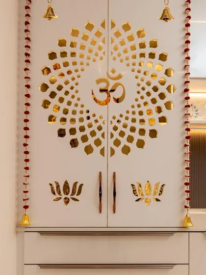 This compact pooja unit is integrated into a larger cabinet, featuring beautiful white doors with a golden "Om" and lotus motif, and hanging bells for a traditional touch.