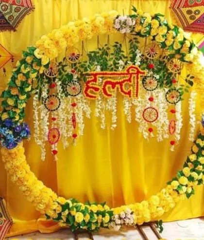 A close-up of a circular Haldi backdrop decorated with yellow flowers and traditional wheel-shaped hangings.