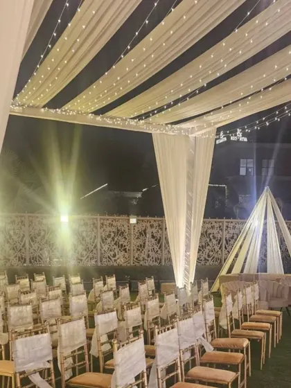 An elegant outdoor ceremony seating arrangement. The white tent is decorated with fairy lights, and the chairs are neatly arranged on the lawn, facing the main stage.