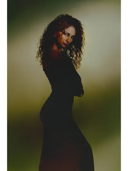A simple, elegant silhouette shot. The model is styled in a classic black dress, and the soft, greenish background light outlines her form and curly hair beautifully.
