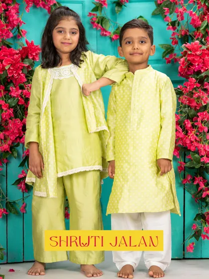 A brother and sister duo in a light lime green set. The boy's kurta has a subtle dot print, while the girl's outfit features a draped kurta with a delicate jacket, creating a fresh and modern festive look.