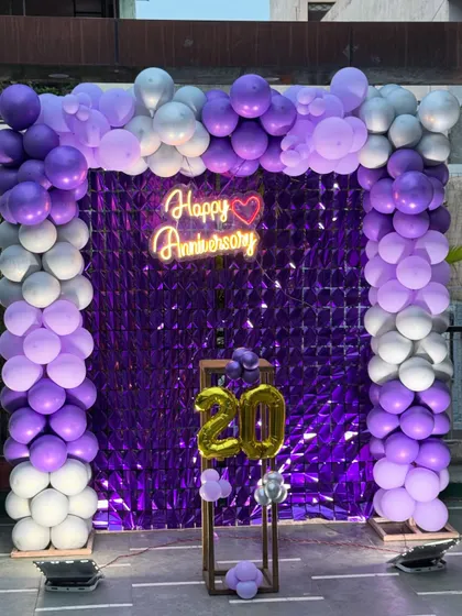 A grand 20th-anniversary celebration with a purple shimmer wall. The entrance is framed by a purple and silver balloon arch, with a neon sign and the number '20' on a pedestal.