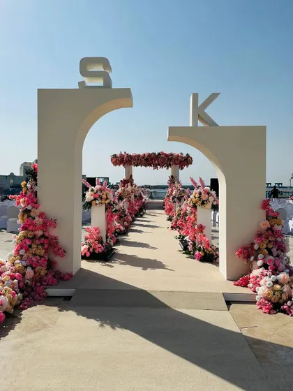 When the entrance archways on your wedding day look this grand and gorgeous. This modern arch, personalized with the couple's initials and adorned with pink flowers, sets the stage for a beautiful beachside ceremony. 🩷