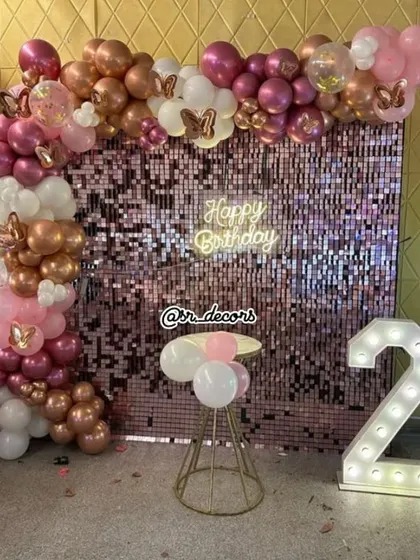 A sweet and simple setup for a second birthday, featuring a pink shimmer wall, a rose gold and white balloon arch, and a large light-up number '2'.