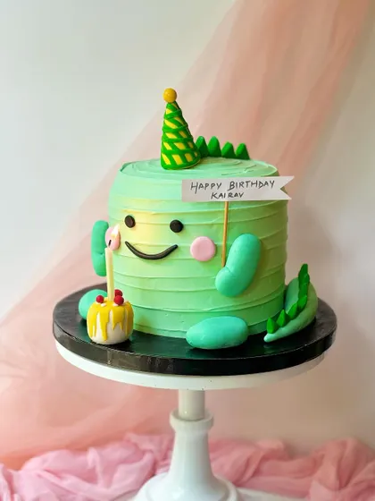 A cute and quirky monster-dinosaur cake for a birthday celebration. The entire cake is designed to be a friendly green monster, complete with a tiny party hat and candle.