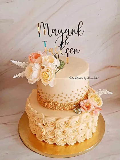 A beautiful two-tier engagement cake that combines different textures. The bottom tier features piped rosettes, while the top is smooth, both tied together with gold details and fresh flowers.