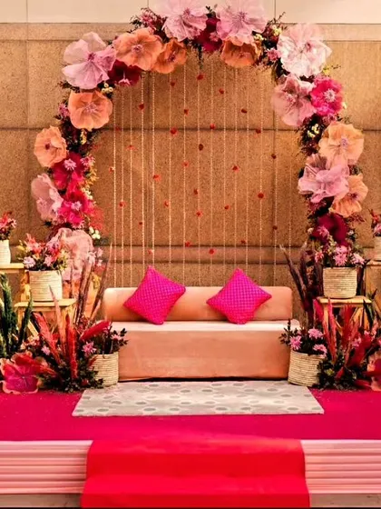 A beautiful indoor Mehendi or Haldi stage decorated with a large arch of paper flowers in shades of pink and peach. The simple sofa and bright pink carpet create a cheerful and elegant space for the ceremony.