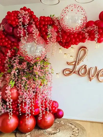A lush and romantic balloon arch in shades of red and pink, intertwined with pink blossoms. The "Love" foil balloon on the wall makes this a perfect backdrop for a Valentine's or anniversary party.